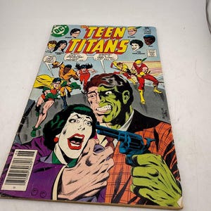 May include: A vintage comic book titled "The Teen Titans" featuring a colorful illustration. The cover art depicts several characters in action, with a dramatic scene of a person pointing a gun. The comic has a DC Comics logo.