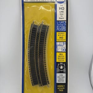 May include: Package of curved HO scale model railroad track pieces. The packaging is blue and yellow, with text that reads "CURVED TRACK" and "READY-TO-USE". The track is black and made by Associated Hobby Manufacturers.