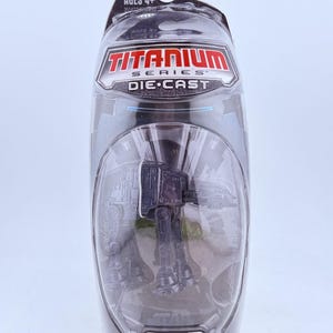 May include: A Star Wars Titanium Series die-cast toy of an AT-AT walker, packaged in a clear plastic blister pack. The packaging features the Star Wars logo and the text "AGES 4+". The toy is dark gray.