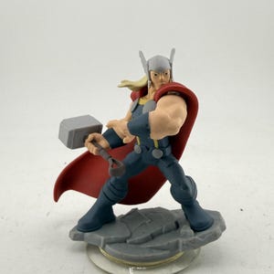 May include: A figurine of Thor, a superhero, in a dynamic pose. He wears a gray helmet, a red cape, and a blue and gray outfit. Thor is holding his hammer, Mjolnir, and stands on a gray platform. The figurine is a collectible item.