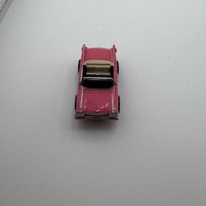 May include: A small, pink toy car with a gold-colored roof and black tires. The miniature vehicle is a classic convertible design, likely from the mid-20th century. The car is set against a plain white background.