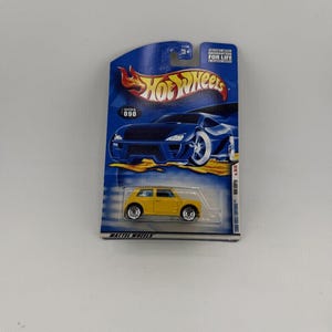 May include: A yellow Hot Wheels die-cast toy car on a blue and white card. The car is a small, detailed model with black wheels. The card features the Hot Wheels logo and the text "2000 First Editions".