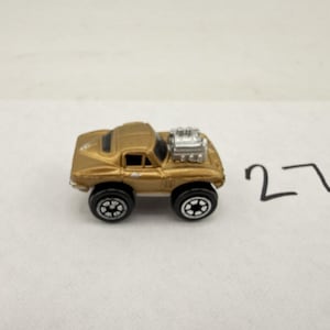 May include: A small, gold-colored toy car with black wheels and a silver engine component. The car has a classic design and is set against a white background. The number "27" is written in black marker.