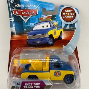 May include: A blue and yellow Race Tow Truck Tom toy from the Disney Pixar Cars series. The toy truck has the text "Race Tow Truck Tom" and "Piston Cup" printed on it. The packaging includes the text "Look! My Eyes Change!"