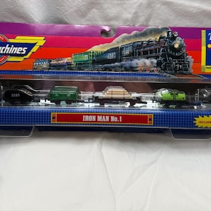 May include: A Micro Machines Iron Man No. 1 train set in original packaging. The set includes a black locomotive, a black tender, a green cargo car, a flatbed car with a tan load, a green tanker, and a gray car. The box features the text Micro Machines.