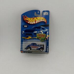 May include: A blue Hot Wheels die-cast model car, a 3-window '34, with an American flag design on the roof and sides. The car is packaged on a blue and white card with the Hot Wheels logo and the text "RACE & WIN ONLINE".