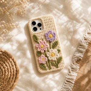 May include: A beige crochet phone case featuring a floral design with three-dimensional flowers in shades of purple, pink, and white, accented with green leaves and yellow centers, covering the back and sides of a smartphone with a triple camera.