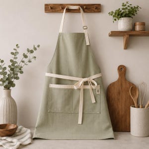 Könnte beinhalten: Eine salbeigrüne Schürze mit cremefarbenen Stoffbändern, einer Schnalle zur Nackenverstellung und zwei Vordertaschen mit cremefarbenem Saum. Die Schürze wird mit einem passenden cremefarbenen Stoffband in der Taille gebunden und hat ein kleines Etikett.
