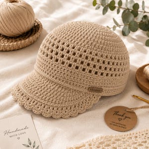 May include: A beige crochet baseball cap featuring a textured lattice pattern on the crown and a scalloped edge along the brim. A small rectangular brown tag with the word Handmade is attached to the side of the cap near the brim.