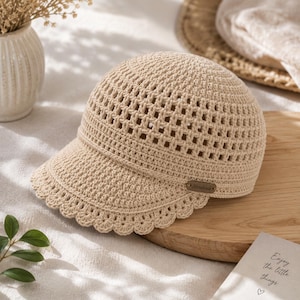 Può includere: Un berretto da baseball all'uncinetto color beige con un motivo a maglia aperta sulla corona e un bordo smerlato lungo la visiera. Una piccola etichetta rettangolare marrone con la parola Handmade è fissata sul lato del berretto.