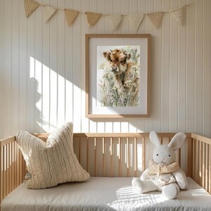 Watercolor highland cow calf printable wall art displayed in natural wood frame above wooden crib in soft neutral farmhouse nursery with knit pillow bunny stuffed animal and pennant banner boho baby room decor