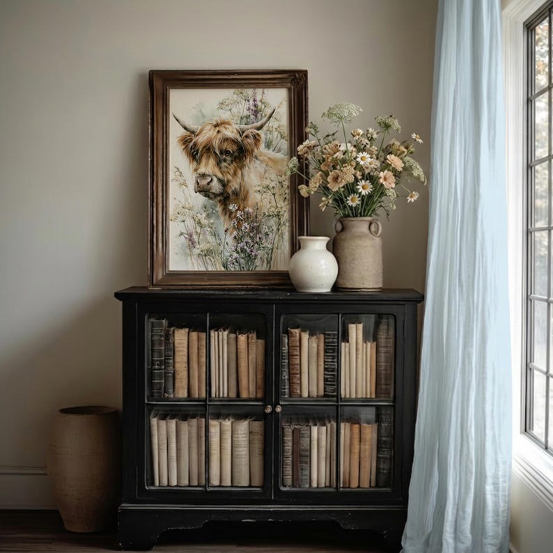 Watercolor highland cow wildflower printable wall art leaning in ornate wood frame on vintage black bookcase with ceramic vases and wildflower stems, cottagecore farmhouse home decor digital download