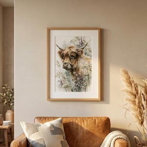 Watercolor highland cow wildflower print displayed in natural wood frame with white mat above tan leather sofa in boho farmhouse living room with pampas grass decor neutral walls digital printable wall art