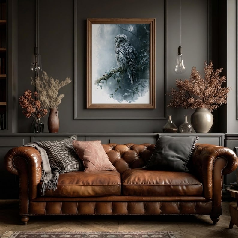 Barred owl watercolor printable wall art displayed in a wood frame above a tufted brown leather Chesterfield sofa in a dark moody living room with dried botanicals and warm Edison bulb lighting
