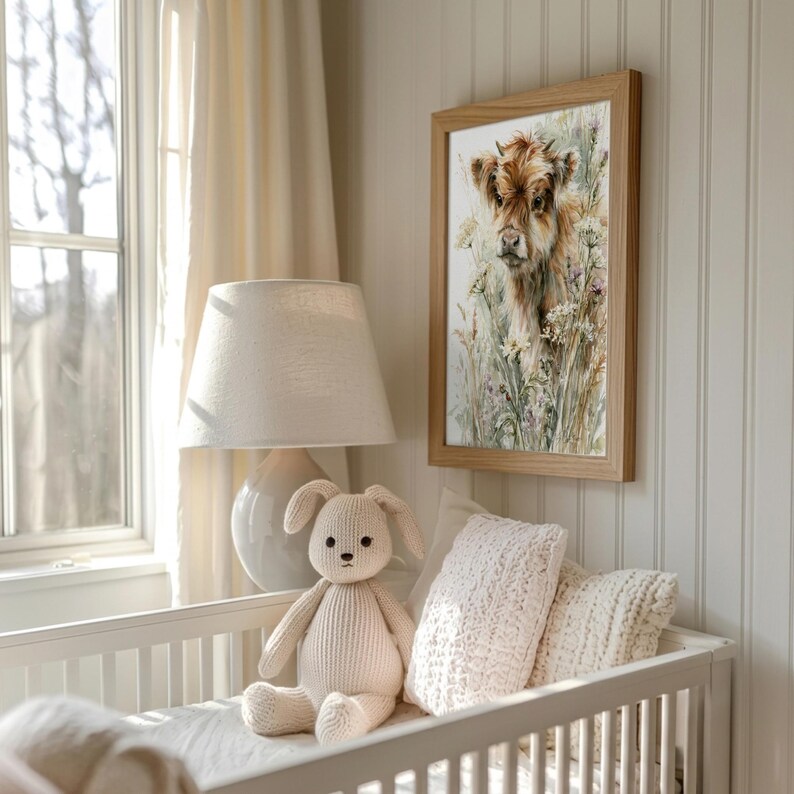 Watercolor highland cow calf nursery wall art in natural wood frame above white crib, styled with knit bunny toy and cream textured pillow in a soft neutral farmhouse baby room