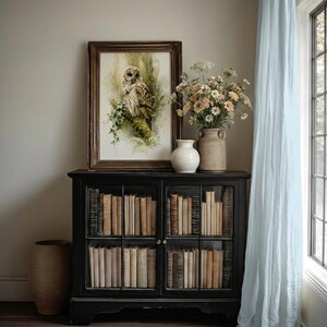 Watercolor owl printable wall art in ornate wood frame leaning on black glass-front bookcase cabinet styled with vintage books, ceramic vases, and wildflowers in a soft cottagecore home library reading nook