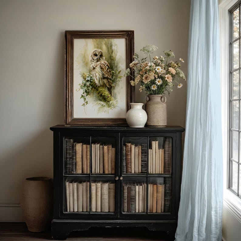Watercolor owl printable wall art in ornate wood frame leaning on black glass-front bookcase cabinet styled with vintage books, ceramic vases, and wildflowers in a soft cottagecore home library reading nook