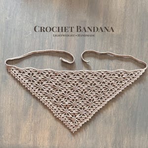 May include: A beige crochet bandana with a triangular shape and a delicate openwork design. The bandana has two long ties for securing around the neck. The text "CROCHET BANDANA LIGHTWEIGHT • HANDMADE" is above the bandana.