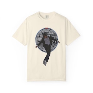 May include: Cream-colored t-shirt featuring a graphic of a person in a black suit and silver shoes, mid-dance pose, inside a blue circle. The circle contains song titles like "Thriller" and "Billie Jean".