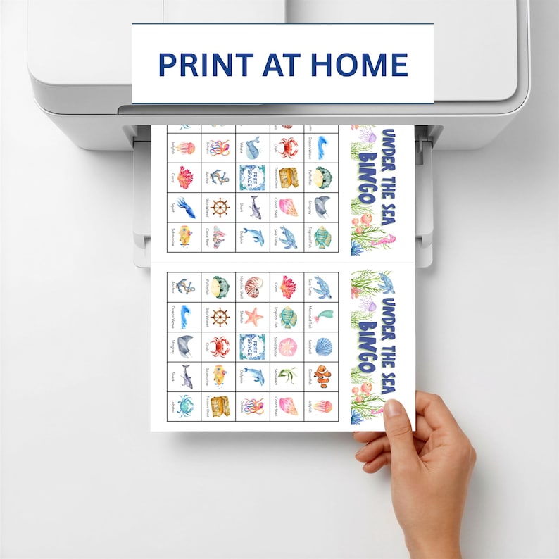 May include: A printer is shown printing two sheets of "Under the Sea Bingo" cards. The cards feature colorful illustrations of marine life, including jellyfish, crabs, and starfish. The text "Print at Home" is displayed on the printer.