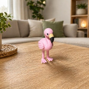 Könnte beinhalten: Eine rosa Flamingo-Figur mit schwarzem Schnabel und gelben Akzenten steht auf einem Holztisch. Der dekorative Vogel ist ein skurriles Akzentstück. Der Hintergrund umfasst ein Sofa, Pflanzen und einen gewebten Untersetzer.