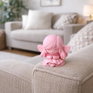Könnte beinhalten: Kleine, rosa Feenfigur mit zarten Flügeln und einem friedlichen Ausdruck. Die Fee kniet mit gefalteten Händen und trägt ein mehrschichtiges Kleid. Die Figur steht auf einer beigefarbenen Sofalehne in einem hellen Wohnzimmer.