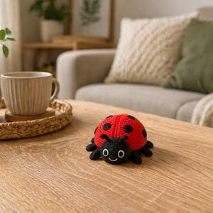 May include: A handmade, red crocheted ladybug with black spots and a black head and legs sits on a wooden table. The ladybug has a smiling face. A vase with greenery and a coffee cup are in the background.