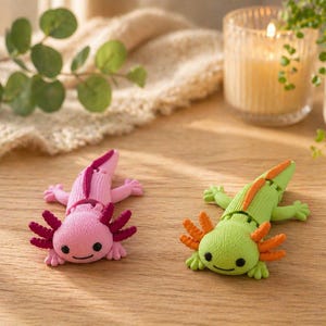 Könnte beinhalten: Zwei farbenfrohe Axolotl-Figuren. Eines ist rosa mit dunkelroten Kiemen und einem dunkelroten Streifen auf dem Rücken. Das andere ist grün mit orangefarbenen Kiemen und einem orangefarbenen Streifen. Beide haben kleine schwarze Augen und liegen auf einer Holzoberfläche.