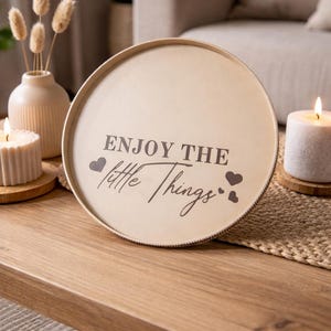 Könnte beinhalten: Ein runder, cremefarbener Tablett mit dem Schriftzug "ENJOY THE little Things" in dunkelgrauer Schrift, verziert mit Herzsymbolen. Das Tablett steht auf einem Holztisch, im Hintergrund Kerzen und eine Vase mit Trockenblumen.