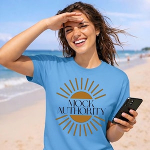 May include: A light blue t-shirt with the text "MOCK AUTHORITY" in black and a sunburst design in brown. The shirt is being worn on a beach.