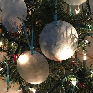 May include: A decorated Christmas tree with several round, silver ornaments. Each ornament is attached to the tree with a light blue ribbon. The tree is lit with white and red lights, creating a festive holiday scene.