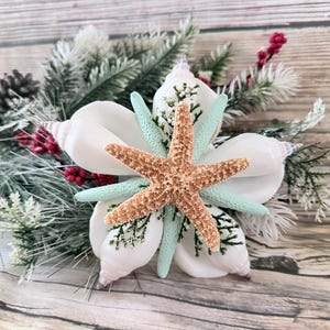 May include: A decorative floral arrangement featuring a large, tan starfish at the center, surrounded by white seashells, light blue starfish, and evergreen branches with red berries. The arrangement is set against a wooden background.