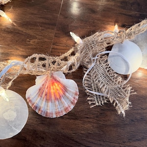 May include: A decorative string of lights with seashells, starfish, and burlap accents. The seashells are illuminated with warm light, and the string is made of natural twine. Perfect for beach-themed decor.