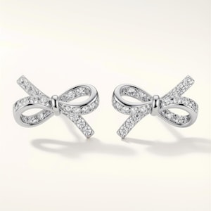 Peut inclure: Une paire de boucles d'oreilles argentées en forme de nœud, chacune incrustée de petites pierres précieuses claires et scintillantes. Les boucles d'oreilles sont présentées sur un fond blanc, mettant en valeur leur design délicat.