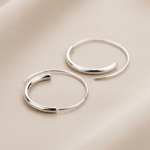 May include: A pair of silver spiral hoop earrings. Each earring features a thicker, rounded section that tapers to a fine wire. The earrings are presented against a neutral, beige backdrop.