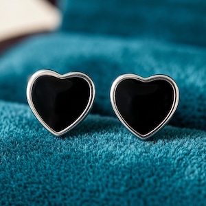 May include: A pair of heart-shaped stud earrings. Each earring has a glossy black heart within a silver-coloured frame. The earrings are displayed on a textured teal surface, creating a striking contrast.
