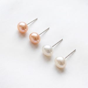 May include: Four stud earrings featuring smooth, round, lustrous pearls attached to silver-toned metal posts. Two of the pearls are a soft peach-pink hue, while the other two are a classic creamy white colour, all arranged in a diagonal line.