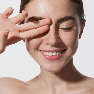 May include: A close-up shot of a person with fair skin, freckles, and dark hair. The person is smiling with their eyes closed, and their hand is covering one eye. The background is plain white.