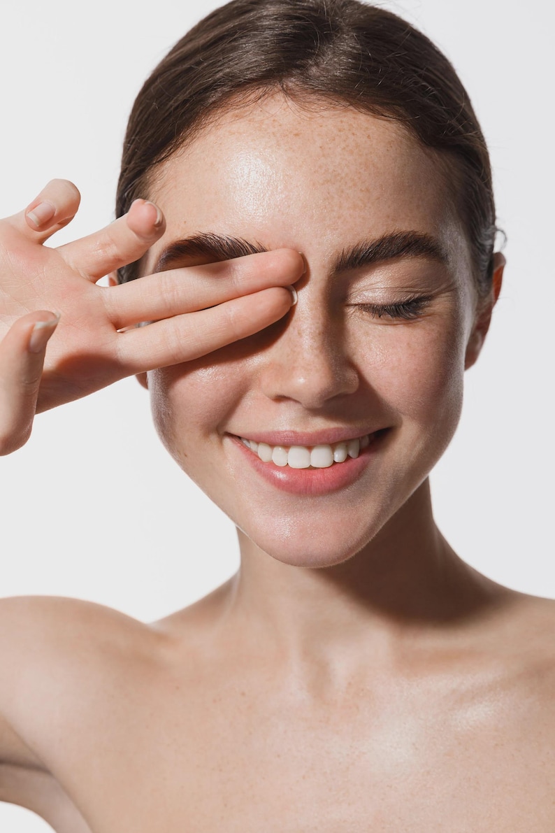 May include: A close-up shot of a person with fair skin, freckles, and dark hair. The person is smiling with their eyes closed, and their hand is covering one eye. The background is plain white.