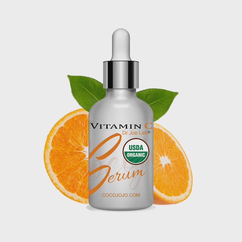 May include: A silver bottle of Vitamin C serum with a dropper, orange slices, and green leaves. The bottle has the text "Vitamin C Serum" and "USDA Organic" on the label. The website address "COCOJOJO.COM" is also visible.