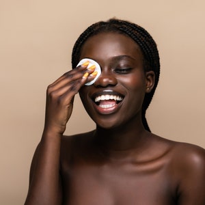 Puede incluir: Una mujer de piel oscura y cabello trenzado sonr&iacute;e mientras usa una almohadilla de algod&oacute;n para quitarse el maquillaje del ojo.