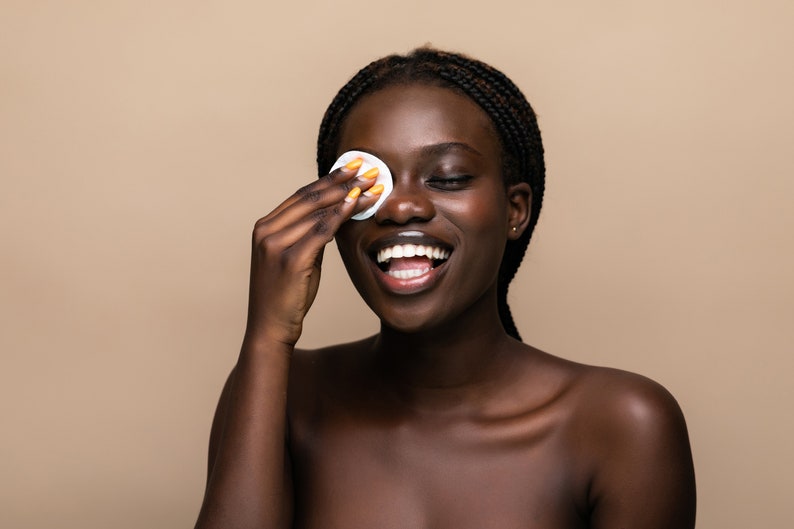 Puede incluir: Una mujer de piel oscura y cabello trenzado sonr&iacute;e mientras usa una almohadilla de algod&oacute;n para quitarse el maquillaje del ojo.