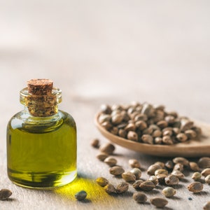 May include: A small glass bottle with a cork stopper filled with green hemp seed oil. A wooden spoon filled with hemp seeds and scattered hemp seeds are on a light wood surface.