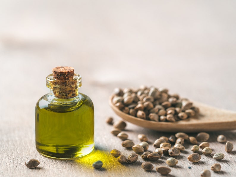 May include: A small glass bottle with a cork stopper filled with green hemp seed oil. A wooden spoon filled with hemp seeds and scattered hemp seeds are on a light wood surface.