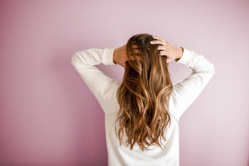 May include: A woman with long, wavy brown hair is wearing a white long-sleeved shirt. She is standing in front of a pink wall and has her hands in her hair.