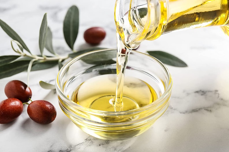 May include: A clear glass bottle pouring golden yellow olive oil into a clear glass bowl on a white marble surface. There are three red berries and green leaves in the background.