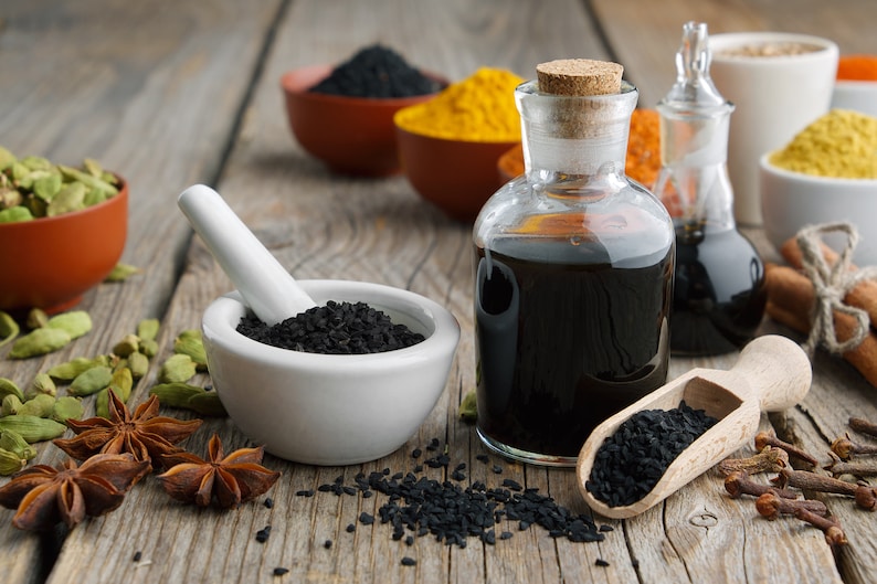 K&ouml;nnte beinhalten: Ein Holztisch mit verschiedenen Gew&uuml;rzen und Kr&auml;utern, darunter schwarze Kreuzk&uuml;mmelsamen, Sternanis, Kardamomkapseln und Zimtstangen. Es gibt auch zwei Glasflaschen gef&uuml;llt mit schwarzem Kreuzk&uuml;mmel&ouml;l und einen wei&szlig;en Keramikm&ouml;rser und St&ouml;&szlig;el mit schwarzen Kreuzk&uuml;mmelsamen darin.