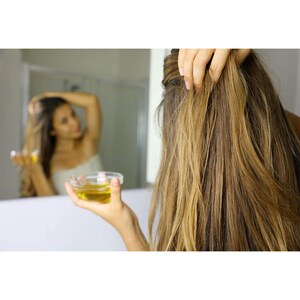 May include: A person holding a small glass bowl of yellow liquid, possibly oil, near long, light brown hair. The person is looking in a mirror, with the reflection showing the person's arm raised. The scene suggests hair care or beauty.