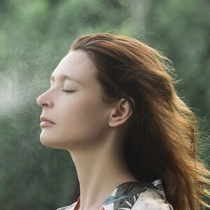 May include: A woman with long brown hair stands in front of a green background, closing her eyes and inhaling a fine mist of water.