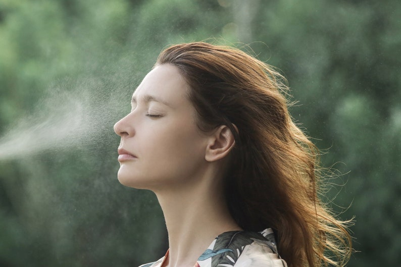 May include: A woman with long brown hair stands in front of a green background, closing her eyes and inhaling a fine mist of water.